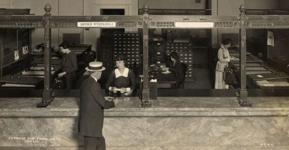 A vintage bank scene with a male customer at the counter, interacting with a female teller. Three women are working behind counters, handling paperwork. Marble counters and decorative ironwork are visible.