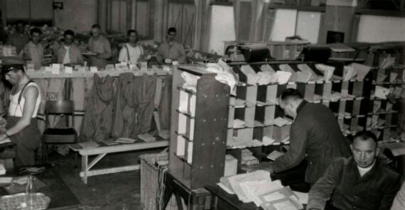 Black and white photo of workers in a busy mailroom, sorting letters into cubbyholes and bags. Papers and envelopes are scattered on desks, with several men focused on organizing the mail.