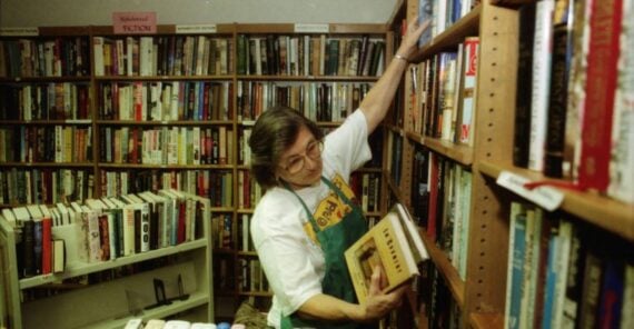 A person wearing glasses and an apron is organizing or shelving books in a bookstore or library, surrounded by bookshelves filled with various books.