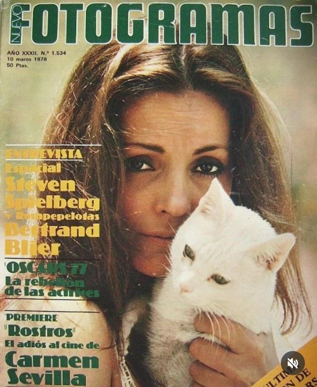 A woman gazes at the camera while holding a white cat. The background features the cover of a Spanish magazine titled "Fotogramas" with text about interviews and film premieres.