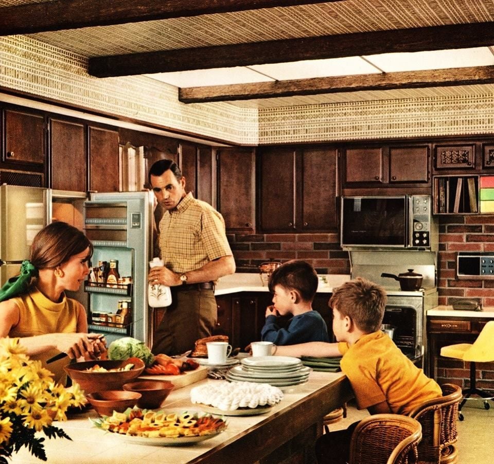 A vintage kitchen scene with a woman and two boys sitting at a counter and a man standing by an open refrigerator. The kitchen has dark wood cabinets, patterned wallpaper, and food on the counter.
