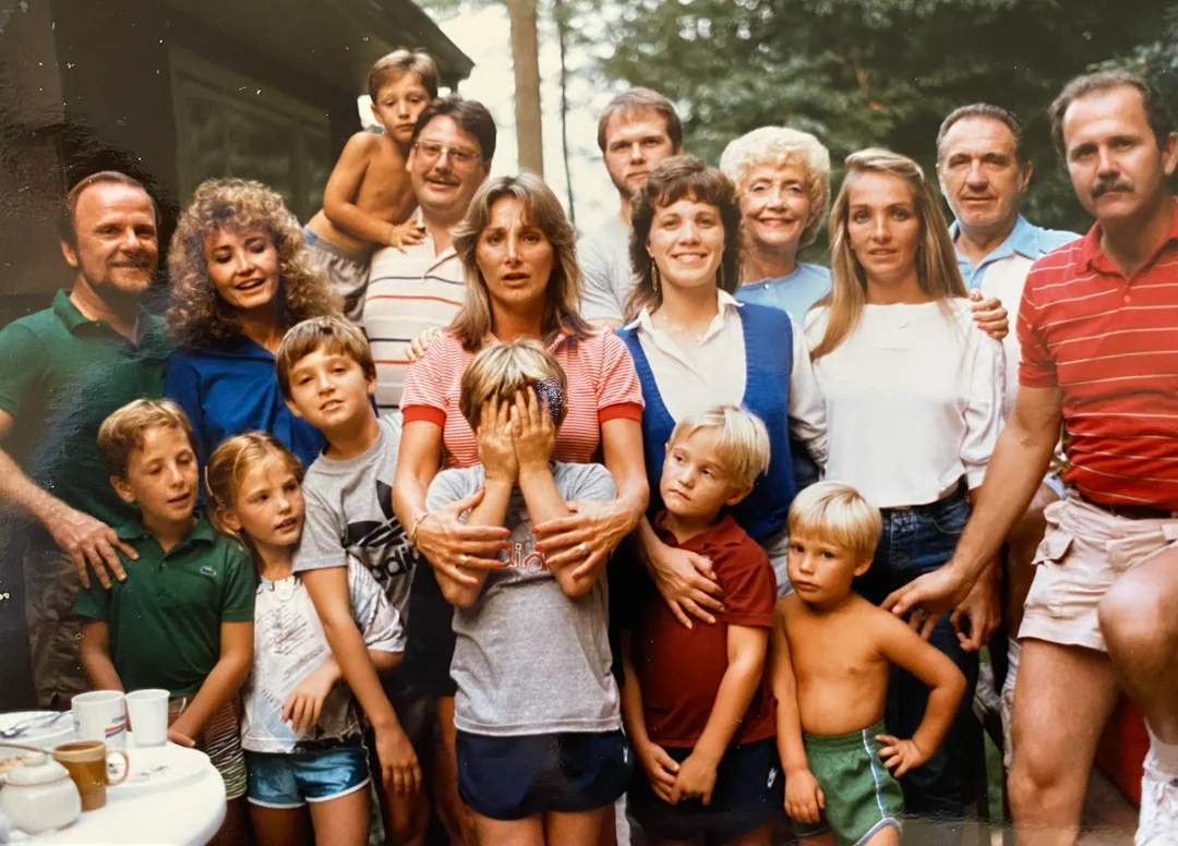 A large family poses outdoors, adults and children standing closely together. One child covers their face while others smile or look at the camera. Trees and a patio are visible in the background.