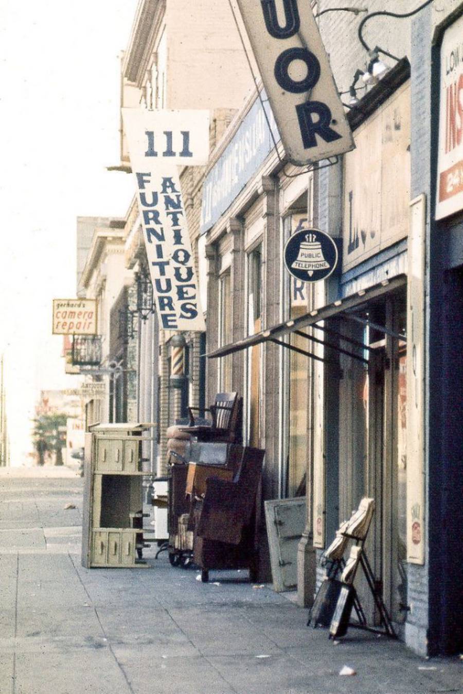 A city sidewalk lined with shops, including antique and liquor stores. Furniture and cabinets are stacked outside, and vertical store signs hang above the storefronts. The street appears empty.