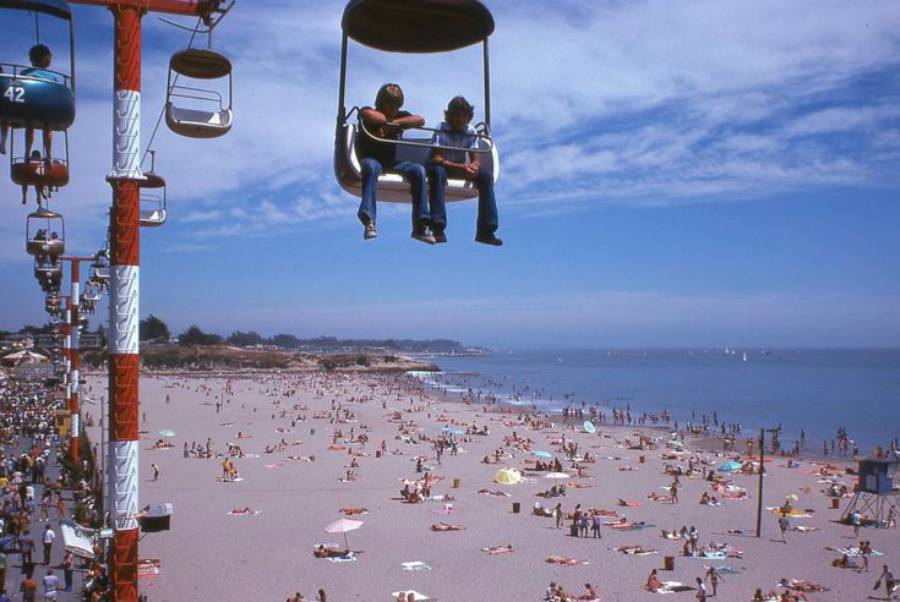 Two people ride a sky lift above a crowded beach on a sunny day. Beachgoers are sunbathing and swimming, with the ocean and a clear blue sky in the background.