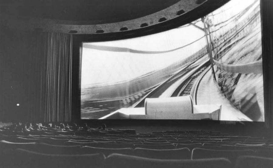 A black-and-white photo of a mostly empty movie theater with a large, curved screen showing a dynamic, first-person view of train tracks. The seats and a few viewers are visible in the foreground.