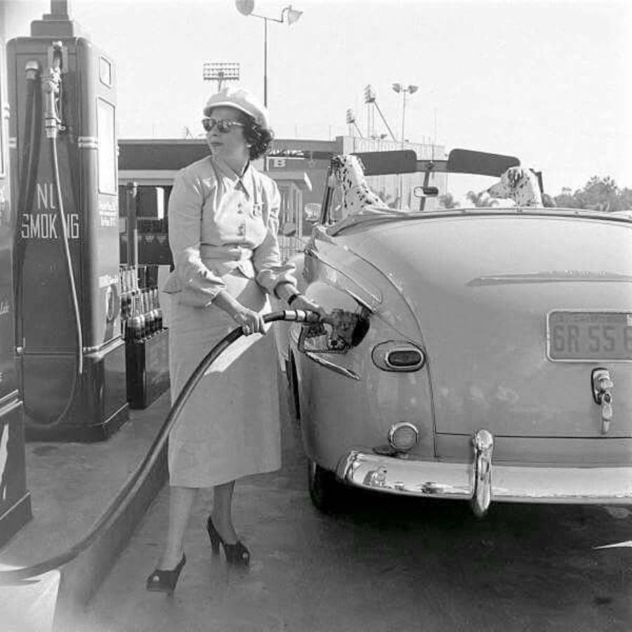 A woman in a skirt suit and sunglasses pumps gas into a vintage convertible at a gas station. The photo appears to be from the mid-20th century.