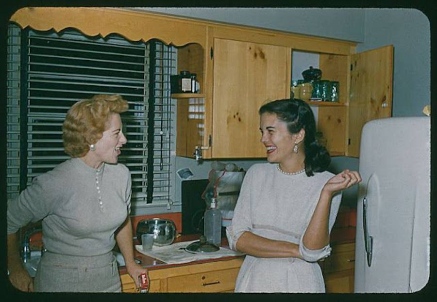Two women stand and smile at each other in a 1950s-style kitchen with wooden cabinets, a white refrigerator, and dishes on the counter. They appear to be engaged in a friendly conversation.
