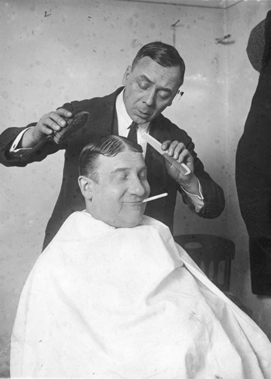 A barber styles a seated man's hair while the man smiles with a cigarette in his mouth. The customer wears a white cape and has his eyes closed, enjoying the moment. Black and white, vintage setting.