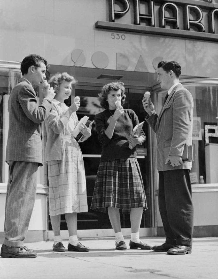 Four young adults in 1940s-style clothing stand outside a pharmacy, eating ice cream cones and holding books, smiling and talking together.