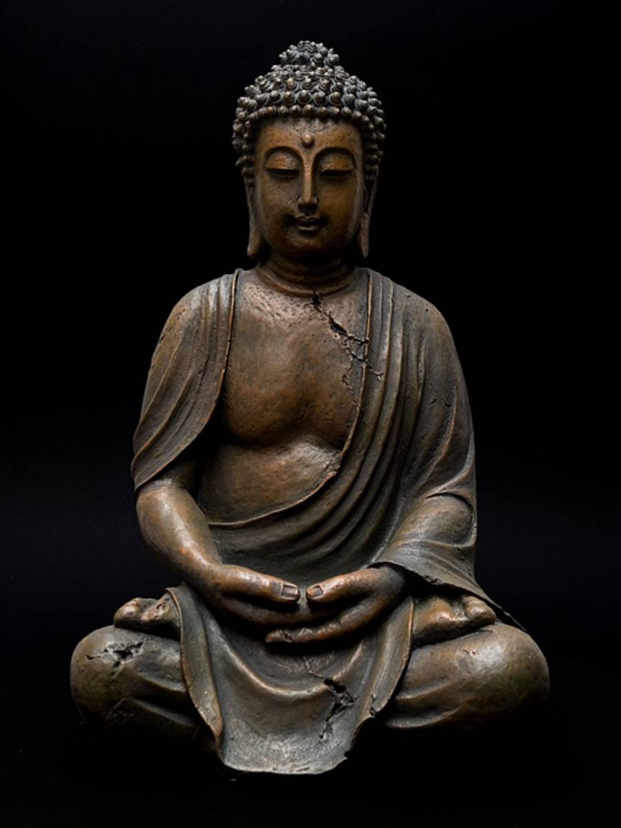 A bronze Buddha statue in a meditative pose with eyes closed, hands resting in its lap, and visible cracks on its surface, set against a solid black background.