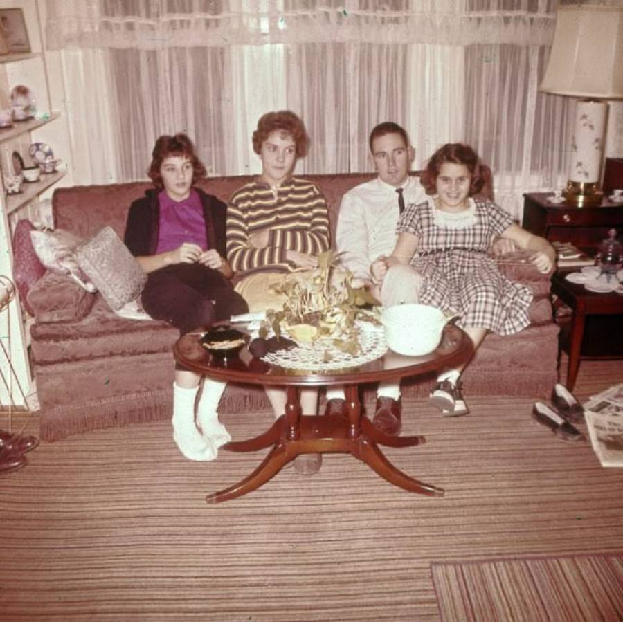 Four young people sit closely together on a vintage sofa in a cozy living room with a coffee table in front of them, lace curtains, and various decorations visible in the background.