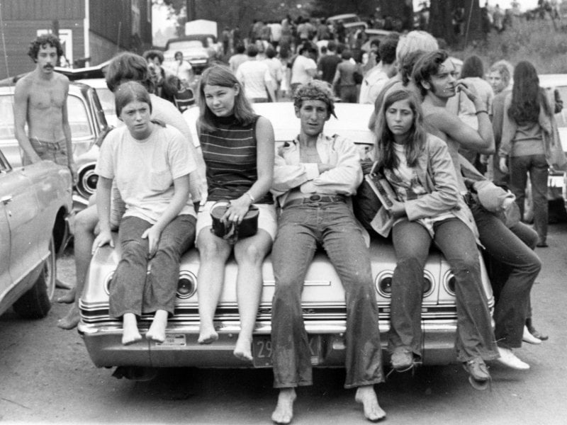 Four young people sit barefoot on the back of a classic car at an outdoor event, with a crowd and parked cars in the background. The scene captures a relaxed, vintage, festival atmosphere.