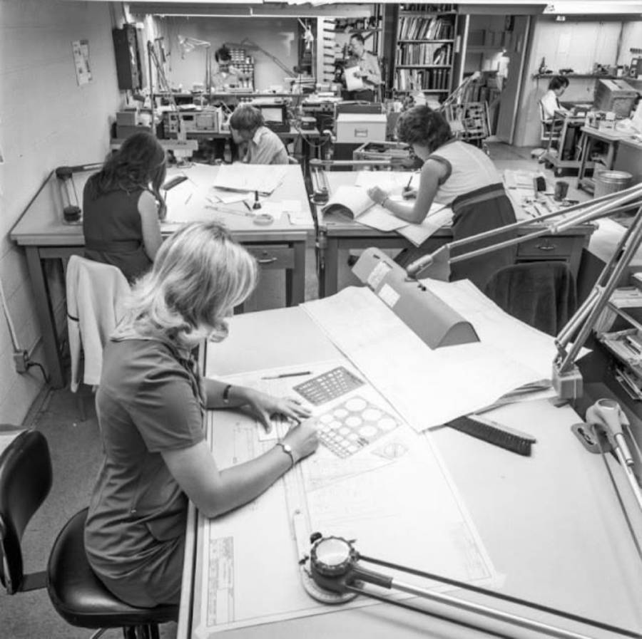 Black-and-white photo of five women working at drafting tables in an office, using paper, rulers, and other drafting tools. The room is filled with desks, equipment, and shelves with documents and books.