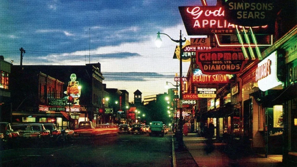 A vibrant city street at dusk, lined with glowing neon signs advertising appliances, beauty salons, and jewelry. Classic cars are parked along the sidewalk, with more vehicles driving under a darkening sky.