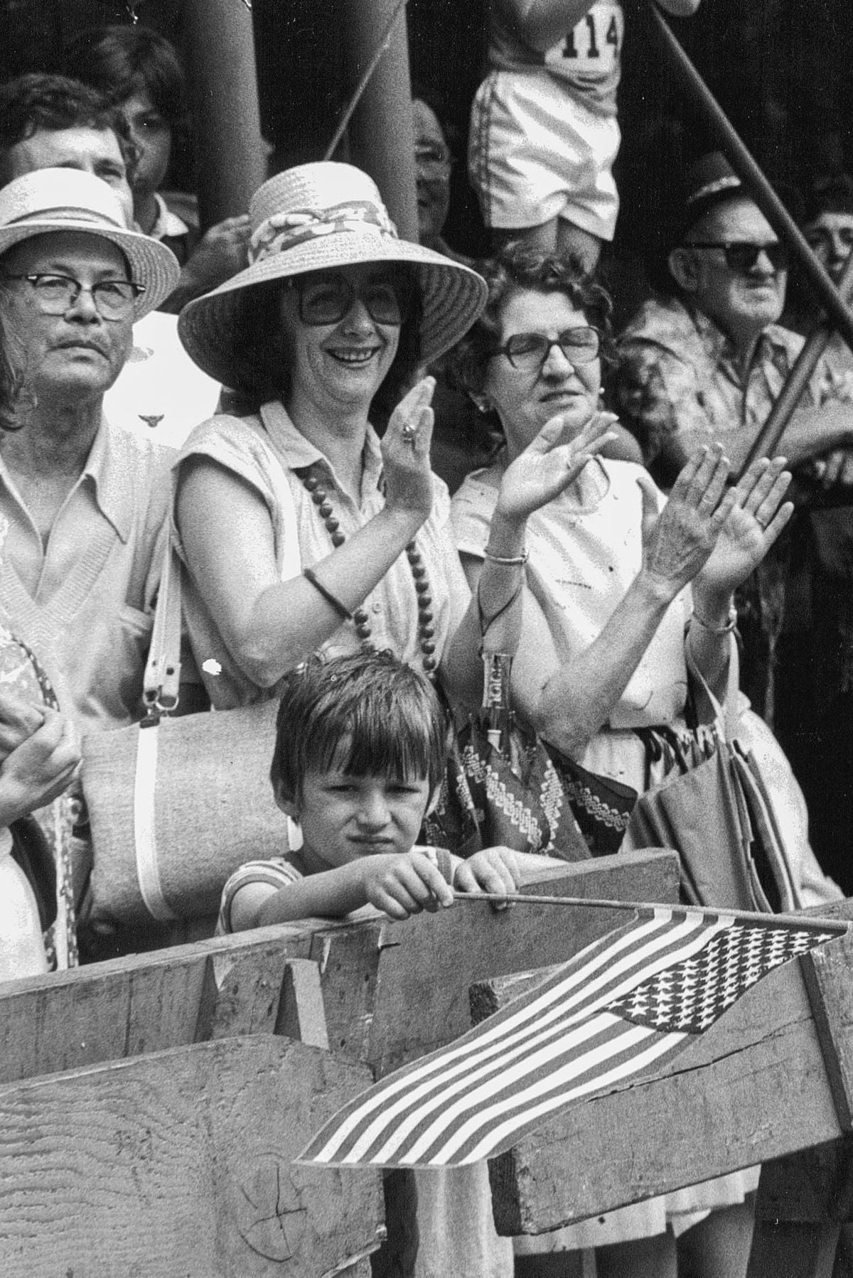 A group of adults clap and smile at an outdoor event while a child stands in front, holding a small American flag and looking at the camera. The scene appears festive and crowded.