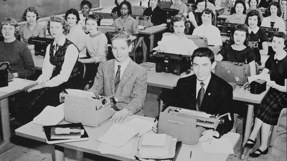 A black-and-white photo of students in a classroom, mostly young women and two young men, seated at desks using typewriters. Papers, books, and typewriters are visible on the desks.