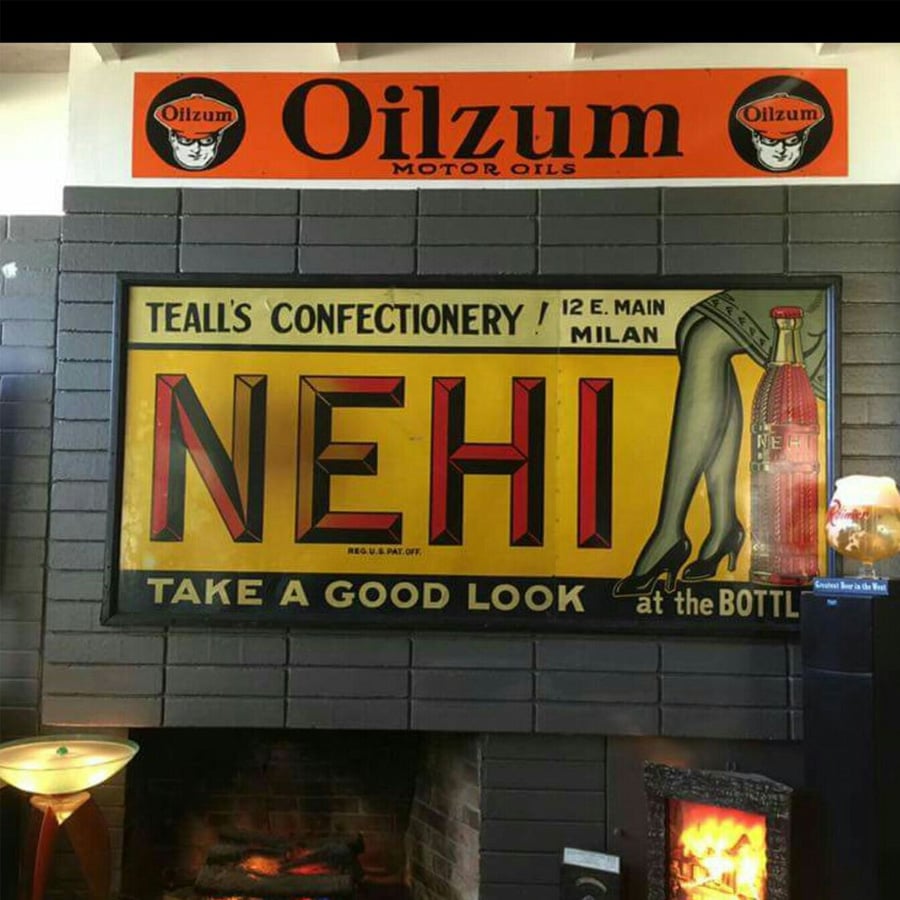 A vintage NEHI soda sign with a woman's legs and a bottle, hangs below an Oilzum Motor Oils sign in a retro-themed room with various decor items.