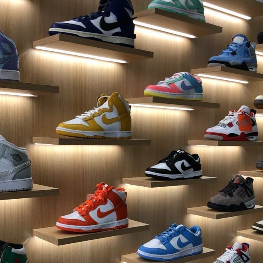 Sneakers are displayed on wooden shelves with built-in lighting. The sneakers come in various colors, including yellow, orange, blue, black, red, turquoise, and white, creating an organized, vibrant wall display.