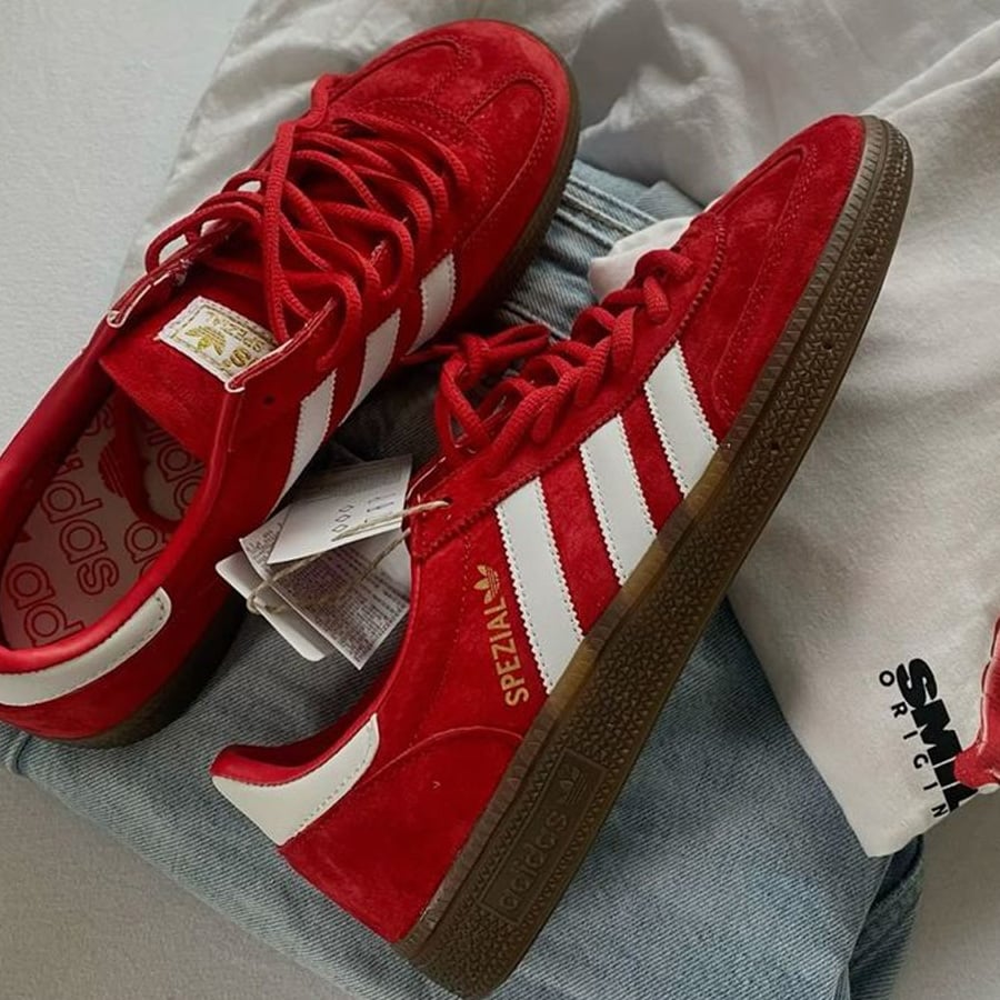 A pair of red Adidas Spezial sneakers with white stripes and brown soles are placed on folded jeans and a white shirt.