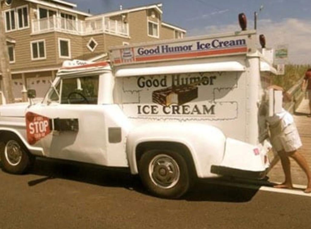 A person stands with their upper body inside the back of a vintage white Good Humor ice cream truck parked on a suburban street, near houses and a sidewalk.