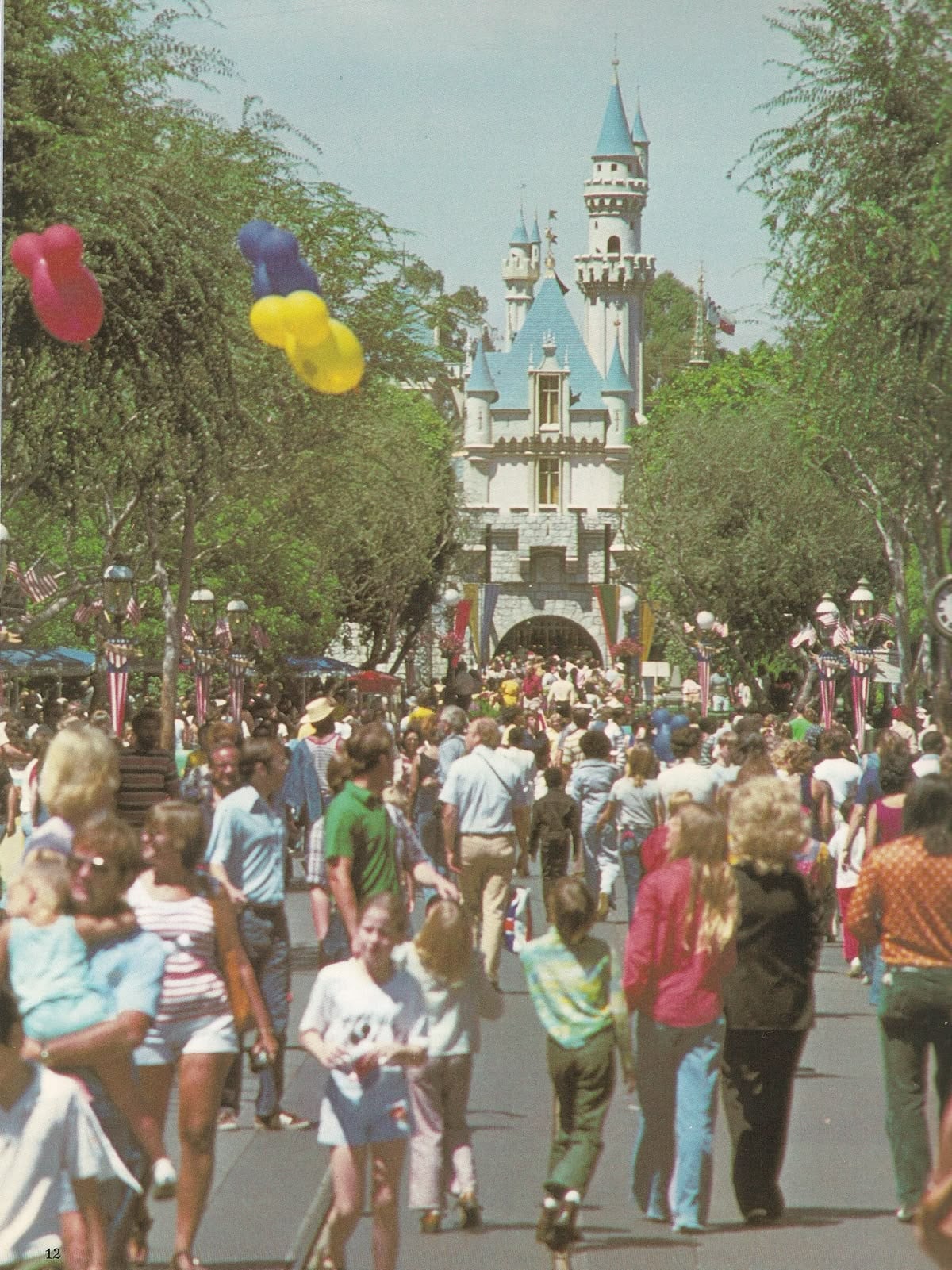 A crowded pathway at Disneyland leads to the Sleeping Beauty Castle, with families, children, and people holding colorful balloons, surrounded by trees on a sunny day.
