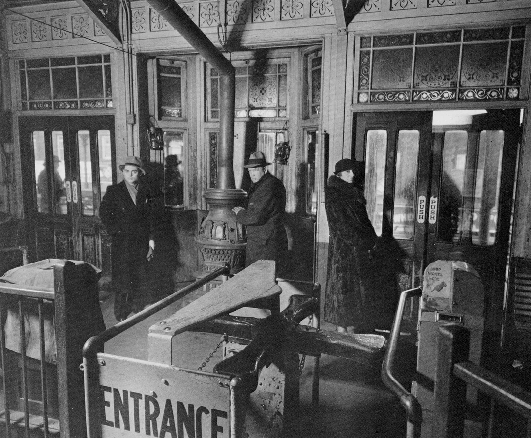 Three people in coats and hats stand in an old subway station near a turnstile labeled "Entrance," surrounded by ornate glass doors and a central stove. The scene is in black and white, suggesting a historical setting.