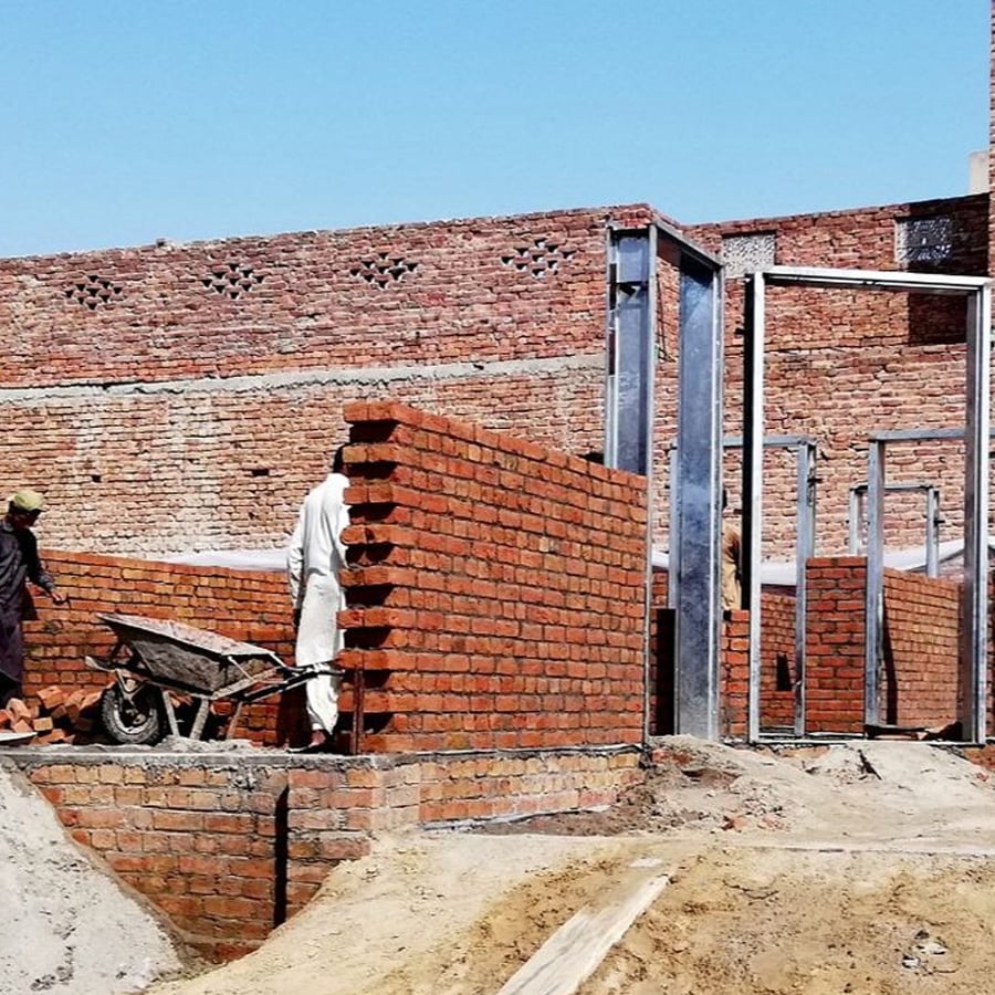 Two construction workers build brick walls at a construction site under a clear blue sky. A wheelbarrow filled with bricks is in front of one of the workers. Metal frames are set up within the structure.