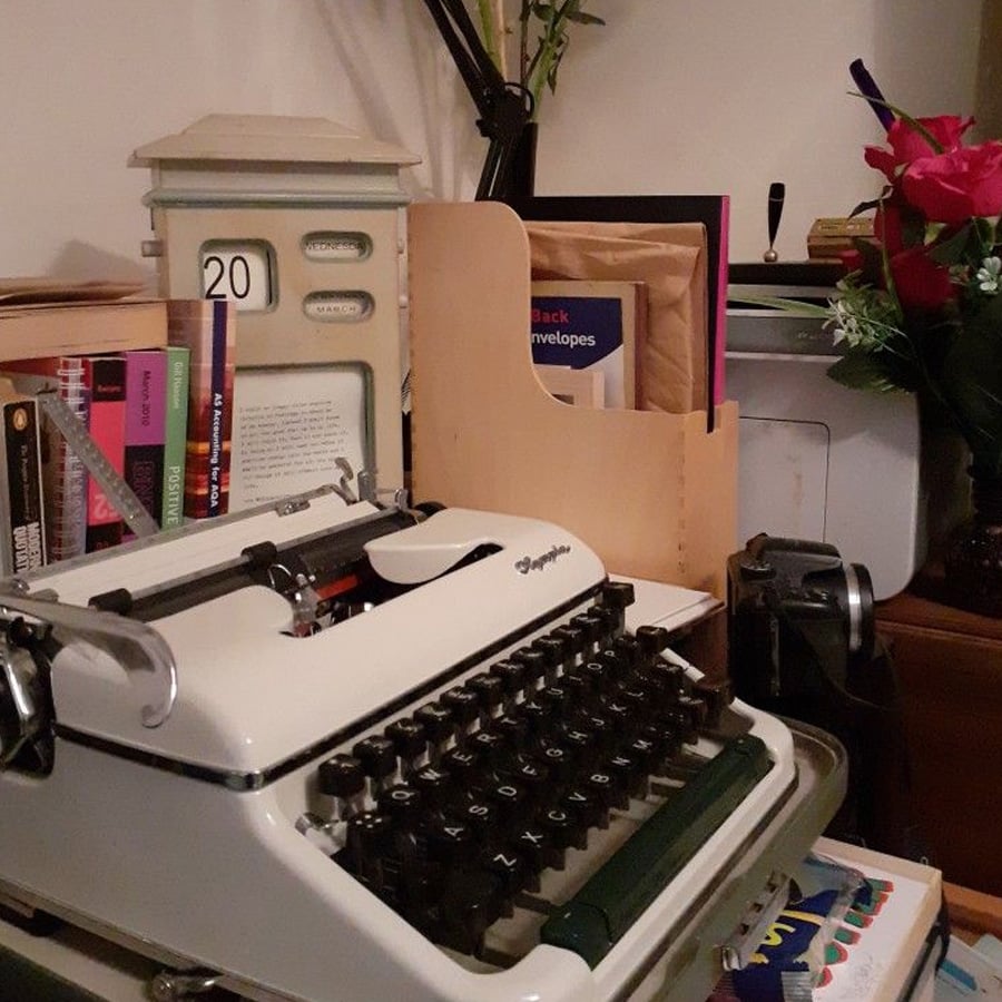 Vintage white typewriter on a cluttered desk with books, a lamp, a calendar showing the number 20, a bouquet of red flowers, and stationery items in the background.