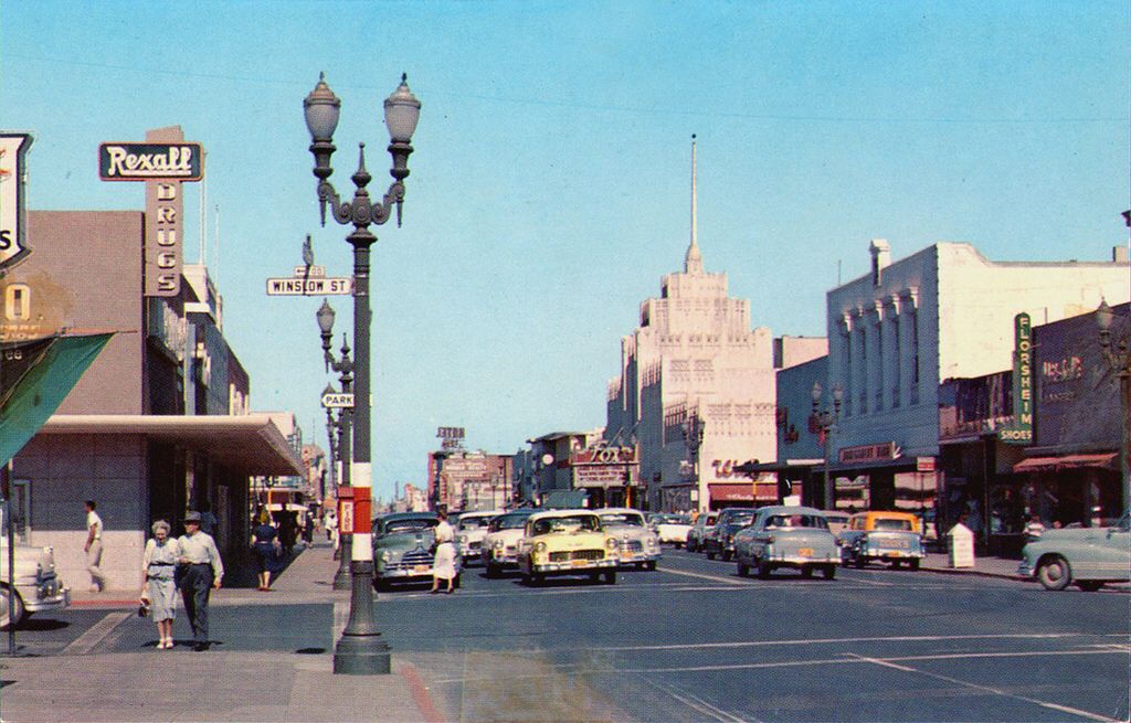 A vintage street scene shows classic cars driving and parked along a busy main street lined with shops, streetlights, and pedestrians. Signs for Rexall Drugs and other businesses are visible under a clear blue sky.