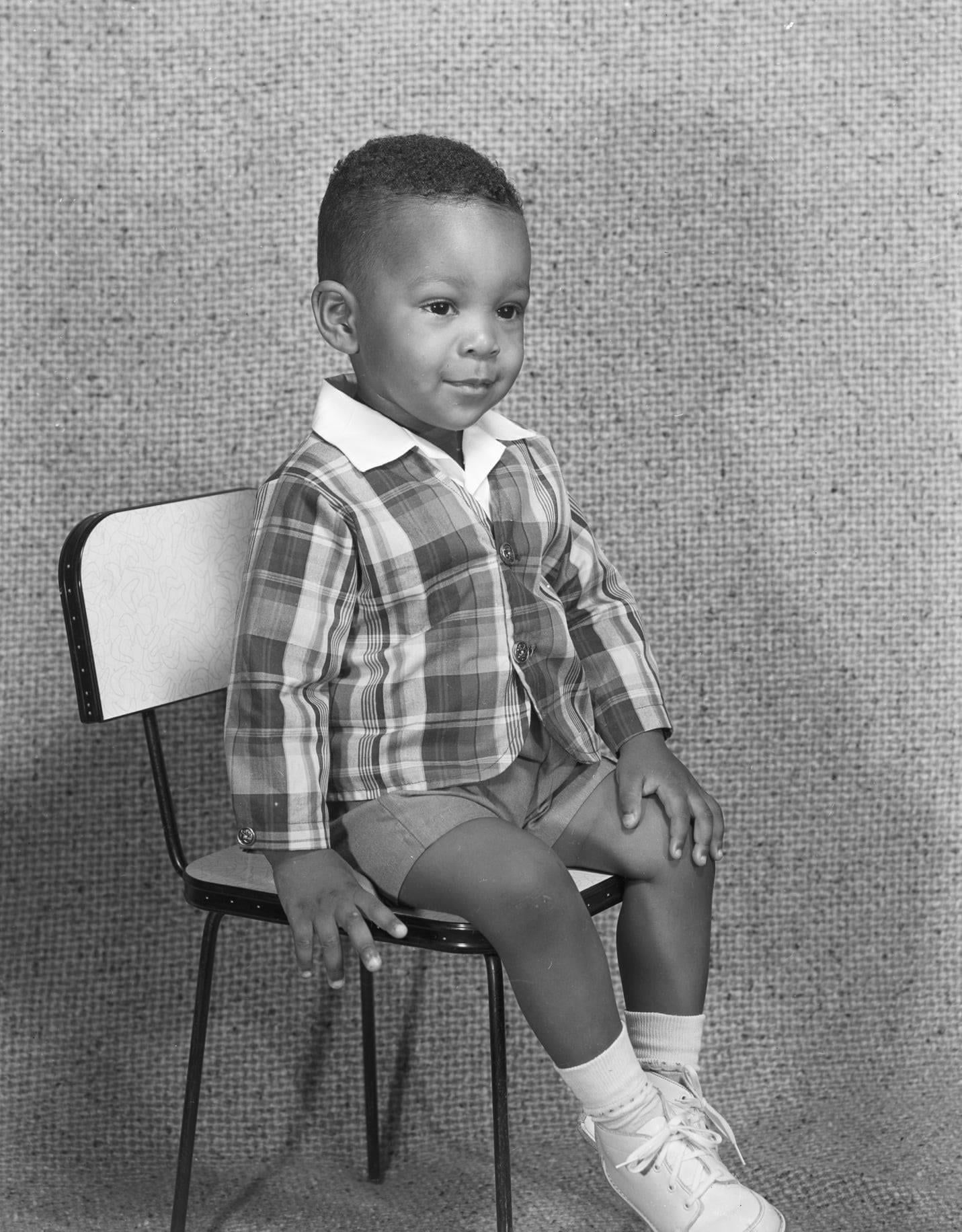 A young child sits on a small chair, wearing a plaid jacket, shorts, socks, and white shoes, posing for a studio portrait against a textured background.