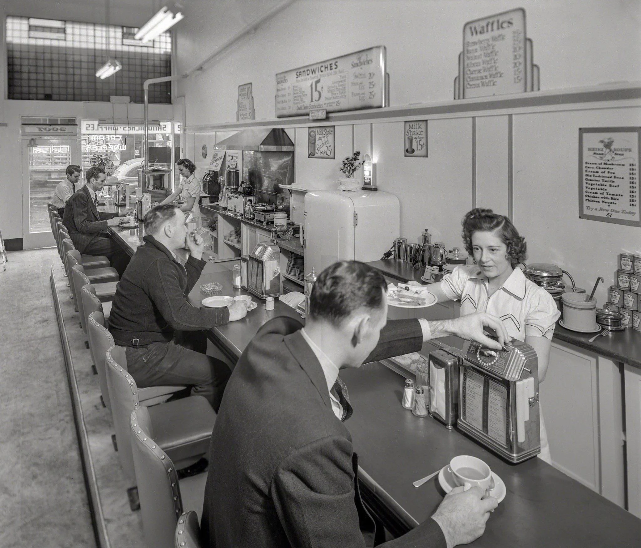 A black-and-white photo of a 1940s diner with a waitress serving coffee to a man at the counter, several patrons eating, and food menus displayed on the wall behind them.