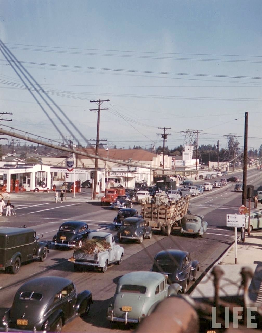 Busy 1950s street scene with vintage cars, utility poles, shops, and a gas station. People walk on the sidewalks, and traffic fills the intersection under clear skies. The image has a LIFE magazine watermark.