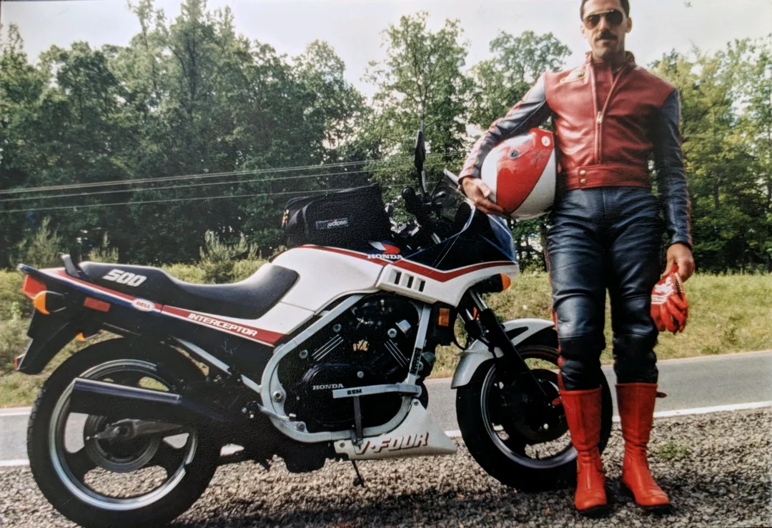 A man in red and black leather gear stands by a white and red Honda VF500F Interceptor motorcycle, holding a matching helmet, beside a road with green trees in the background.