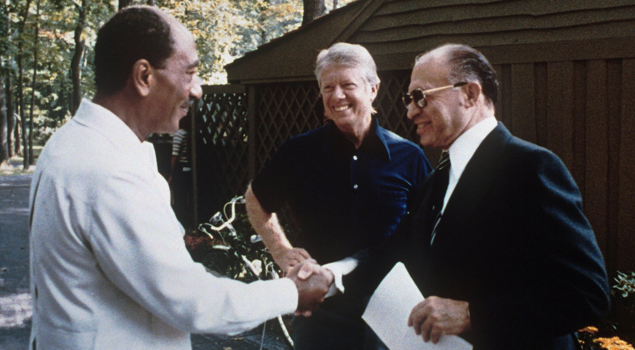 Three men stand outside smiling; two shake hands while the third stands between them, watching. Trees and a wooden building are in the background. All three men appear to be engaged in a friendly conversation.