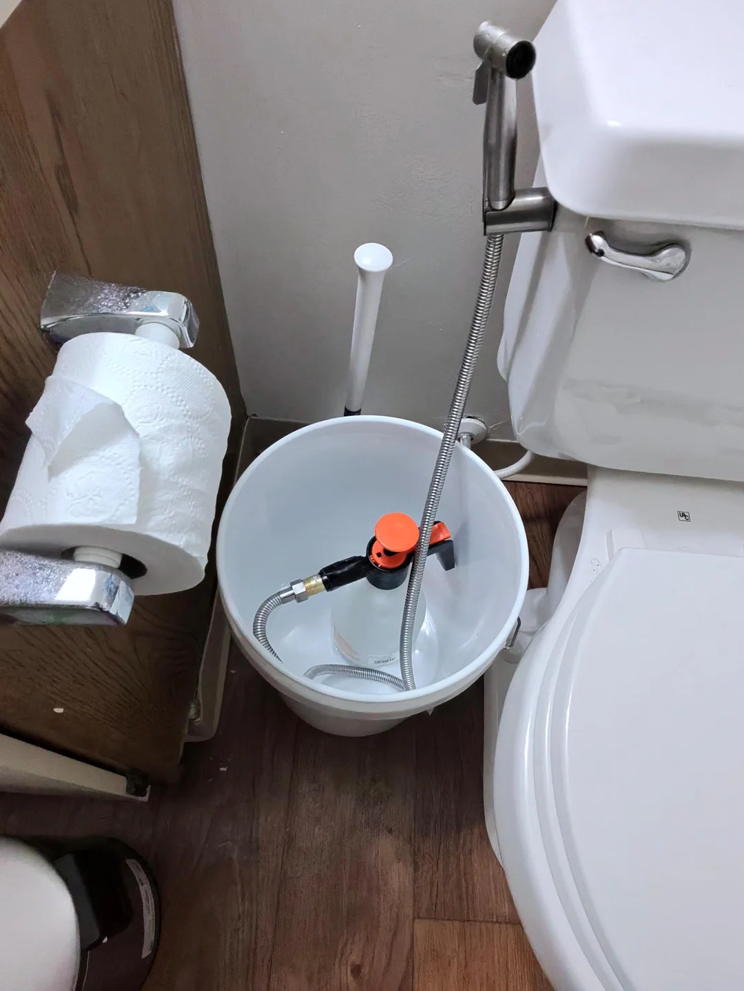 A white bucket with hoses and a valve sits between a toilet and a wall with a toilet paper holder; the bucket appears to be used for plumbing or cleaning purposes.