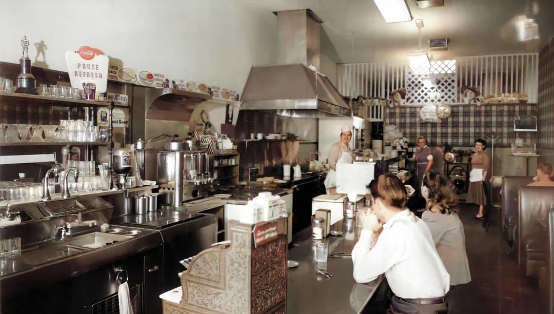 A vintage diner with a long counter, stools, and people seated. Staff work behind the counter, with kitchen equipment and shelves lined with glasses and condiments visible. The scene suggests a busy, classic American eatery.