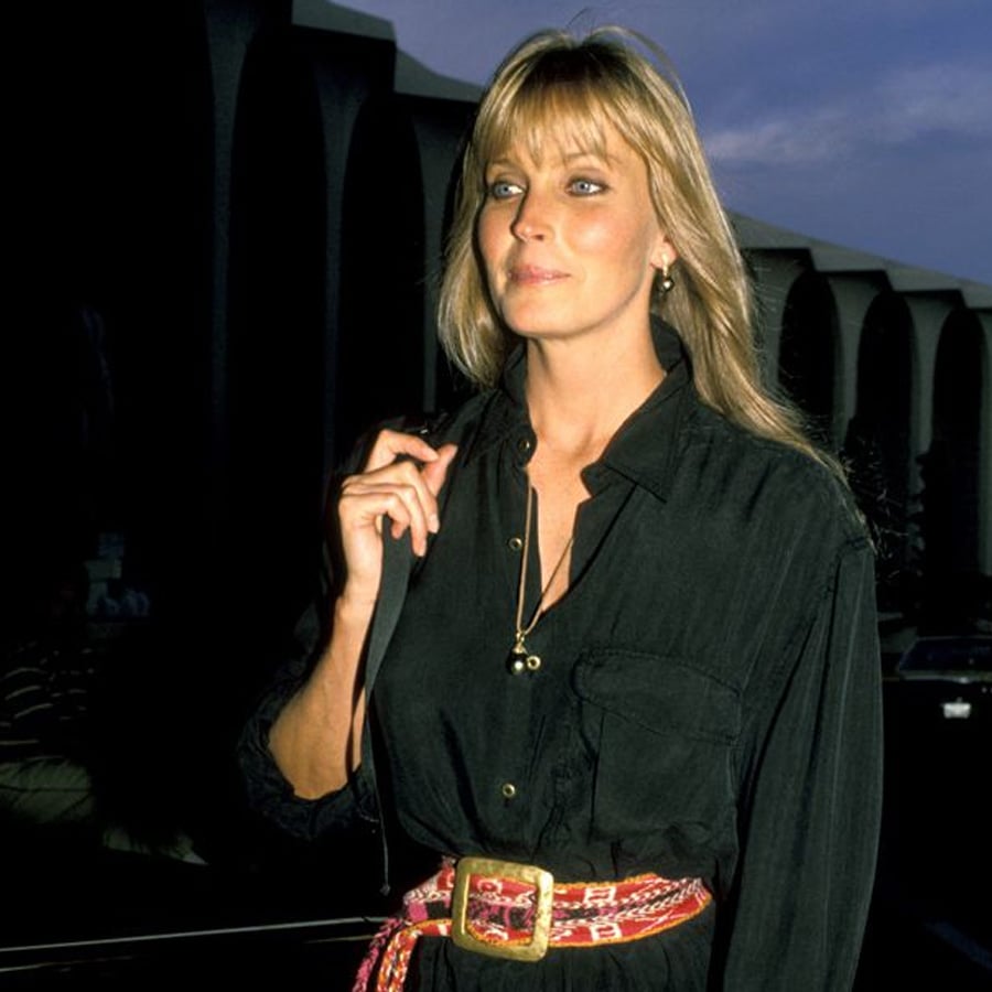 A woman with long blonde hair wearing a black shirt and a colorful belt stands outside at dusk, holding a strap over her shoulder, with a building and dark shadows in the background.