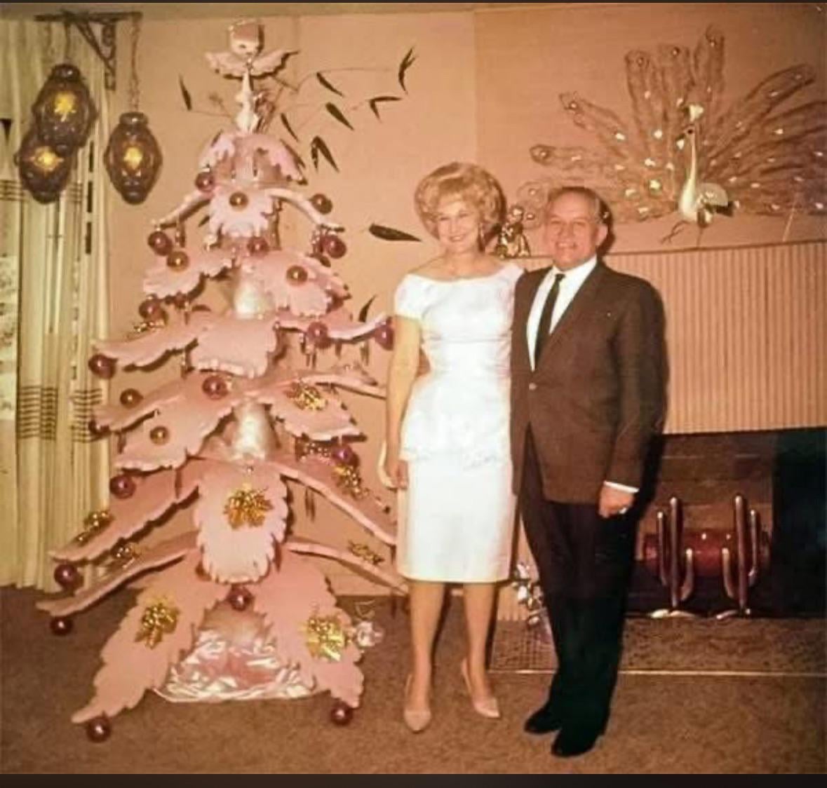 A woman in a white dress and a man in a suit stand by a pink Christmas tree in a retro living room with ornate wall art, including a large peacock decoration.
