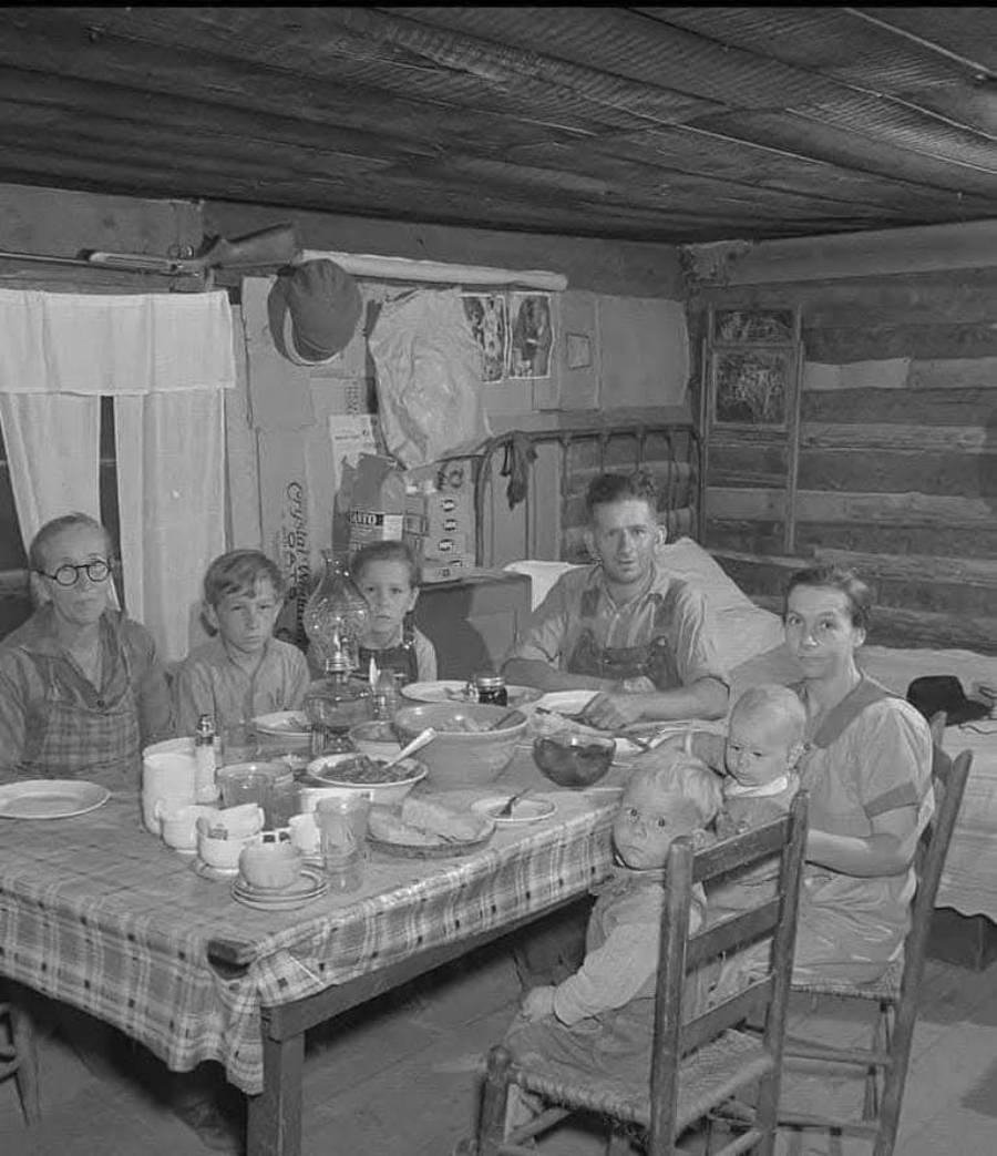 A black-and-white photo of a family of seven sitting around a table in a rustic, wooden cabin, sharing a meal. The table is covered with a checked cloth and simple dishes. The room features wooden walls and household items.