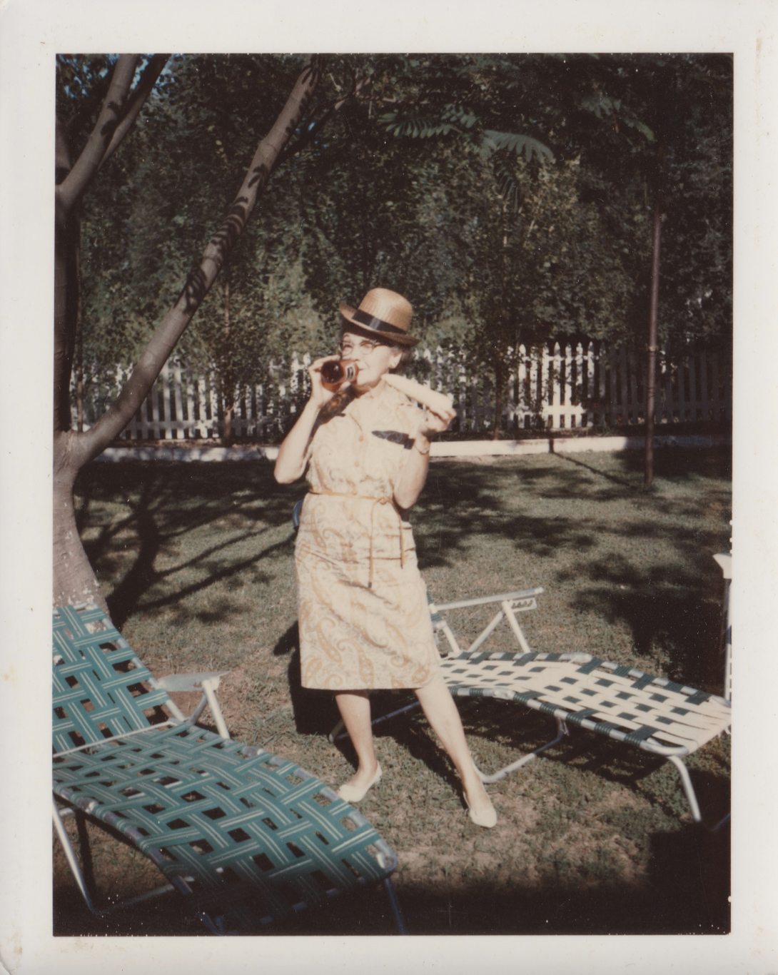 A woman in a patterned dress and hat stands outdoors by lawn chairs, holding a bottle and a sandwich, with trees and a white fence in the background.