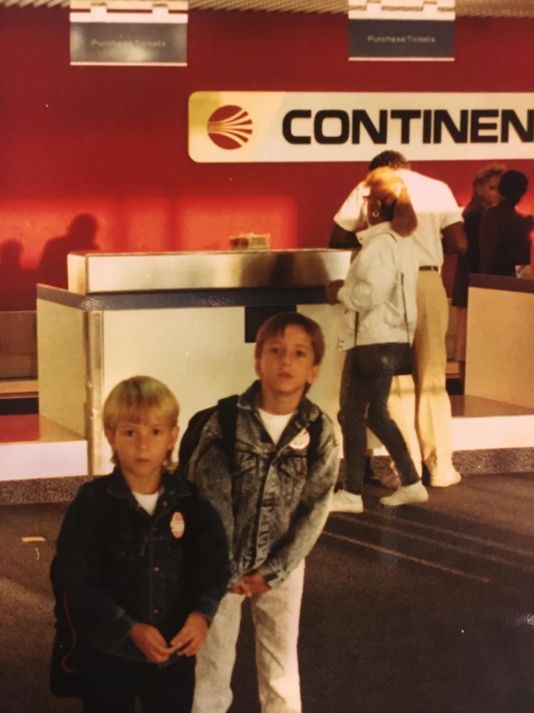 Two young boys with backpacks stand in front of a Continental Airlines counter at an airport. One wears a denim jacket, the other a dark sweater. People are at the counter in the background. The setting has a red wall and airline signage.