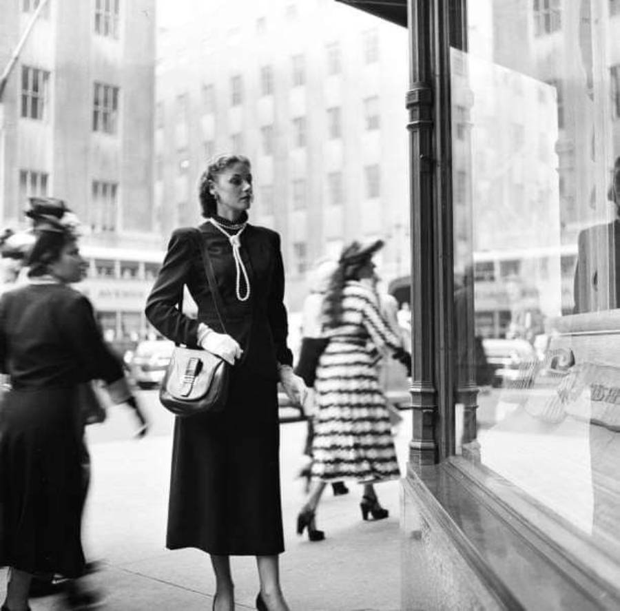 A woman in 1940s attire stands on a city sidewalk, looking into a shop window. She wears a long dark dress, gloves, and carries a handbag. Other women in period clothing walk by in the background.