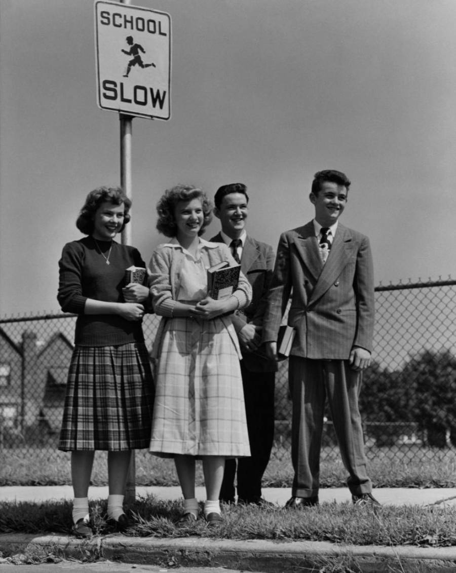Four teenagers in 1950s-style clothes stand smiling near a "SCHOOL SLOW" sign and a chain-link fence, holding books. Two girls wear skirts, and two boys wear suits.