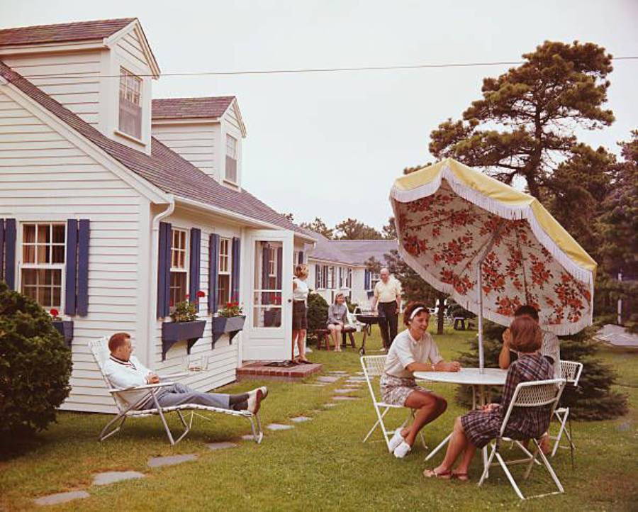 People relax outside a white house with blue shutters; some sit at a table under a floral umbrella, others stand or lounge nearby. The lawn is green with trees and shrubs, creating a casual, summery gathering scene.