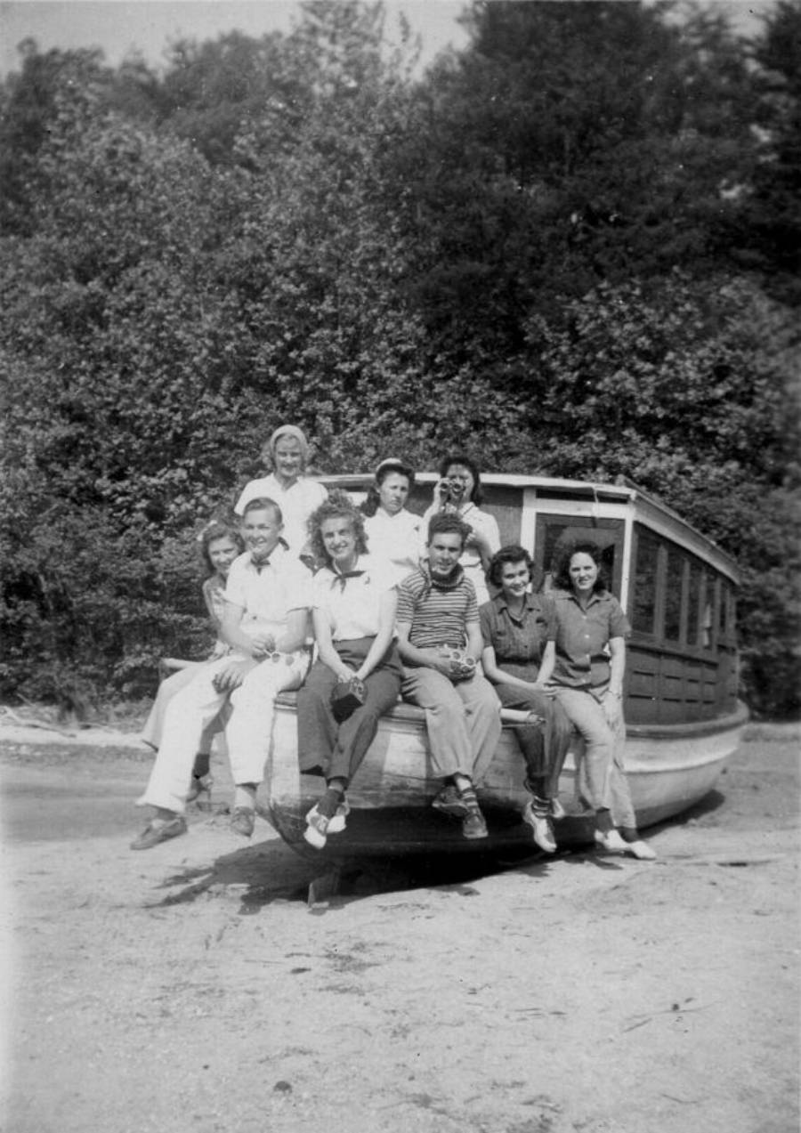 A group of nine young people pose together, sitting and standing on the front of a wooden boat beached on sand, with trees in the background. The photo is black and white.