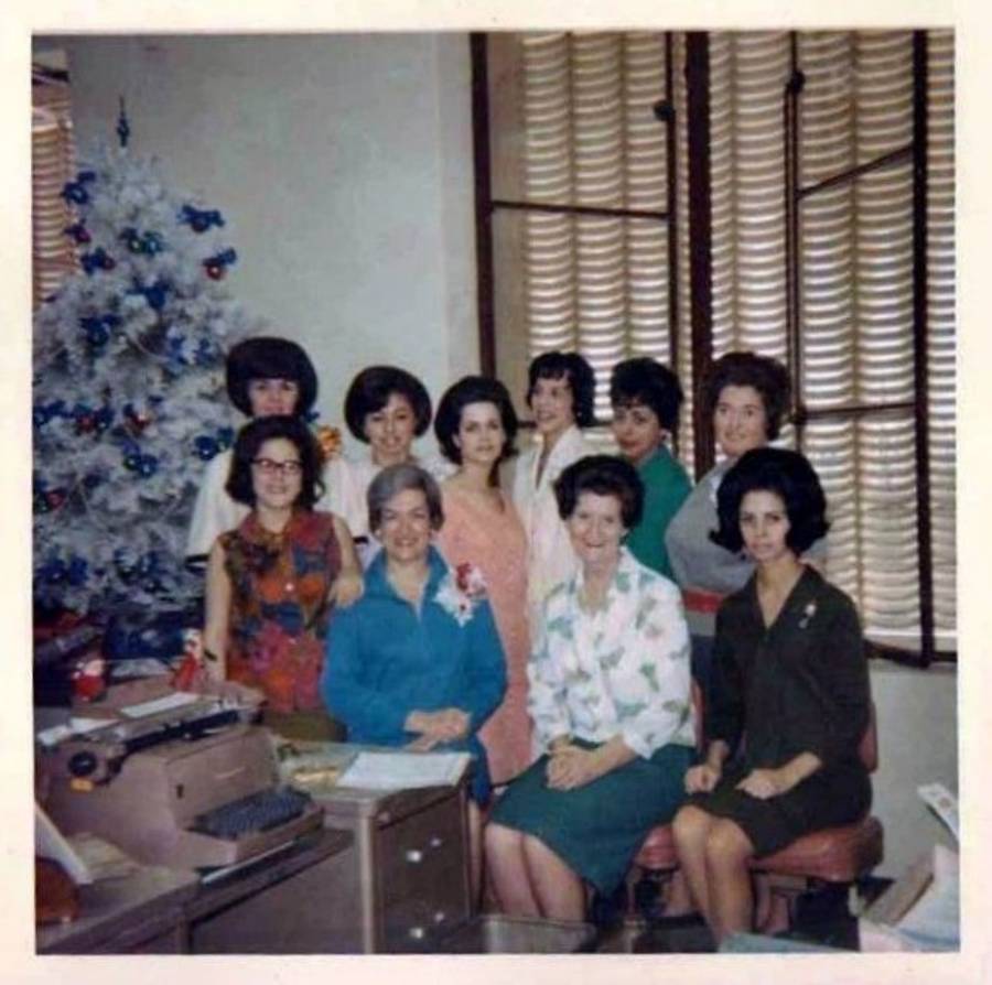 Nine women, dressed in 1960s attire, pose and smile in an office next to a decorated white Christmas tree. Blinds cover the windows, and a typewriter and papers sit on the desk in the foreground.