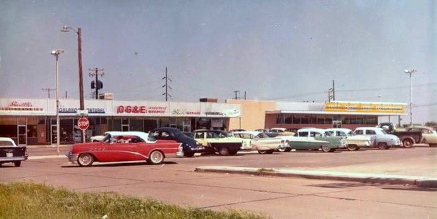 A vintage photo of a 1950s or 1960s shopping plaza with classic cars parked and driving by storefronts, including a grocery store and retail shops under a sunny blue sky.