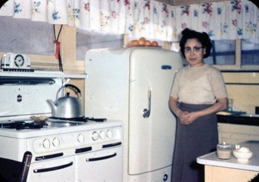 A woman stands in a retro kitchen beside a white refrigerator and stove with a kettle. There are bowls on the counter and floral curtains on the window.