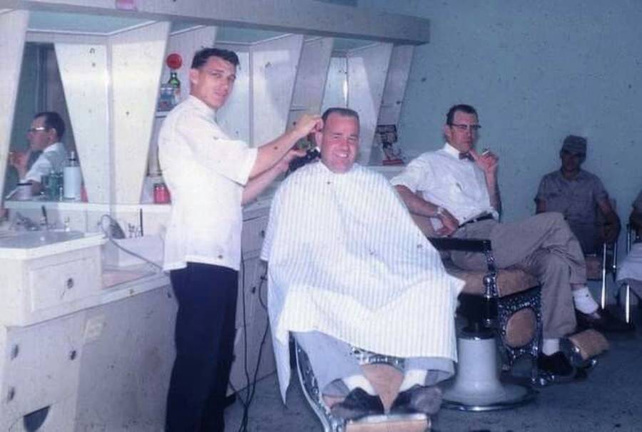 A barber is cutting a smiling man's hair, while another man waits in a chair nearby. The setting appears to be a vintage barbershop with mirrors and retro furnishings.