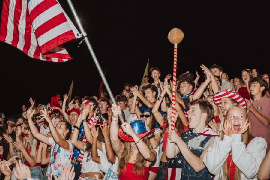 A large group of excited young people celebrate at night, waving an American flag and wearing red, white, and blue clothing and face paint. Many are cheering with their hands up and smiling.